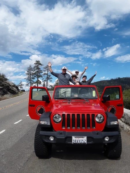 A group of riders gathered in front of a red Jeep at a Sierra mountain overlook.